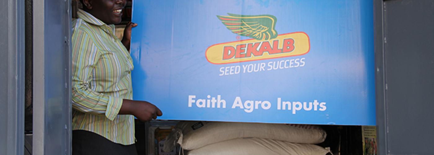 Photo: Woman holds up sign for Dekalb Seed for Success Agro Inputs above bags of grain. 