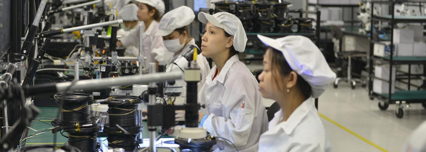 A row of factory workers wear white at a row of workstations. 