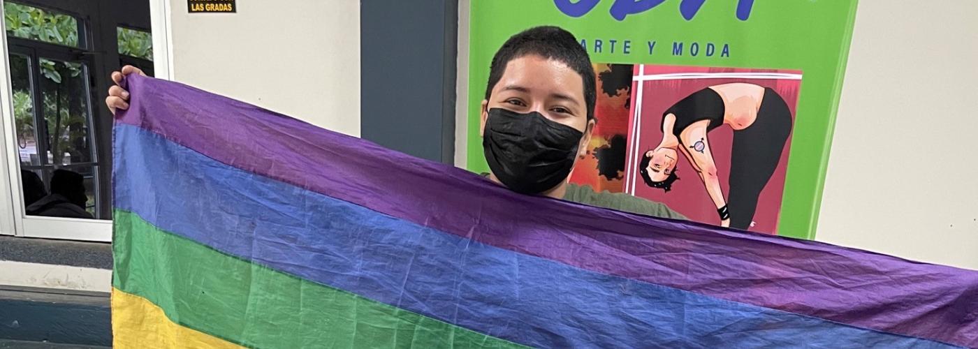 A young man holds up a rainbow flag and stands behind a table filled with artwork 