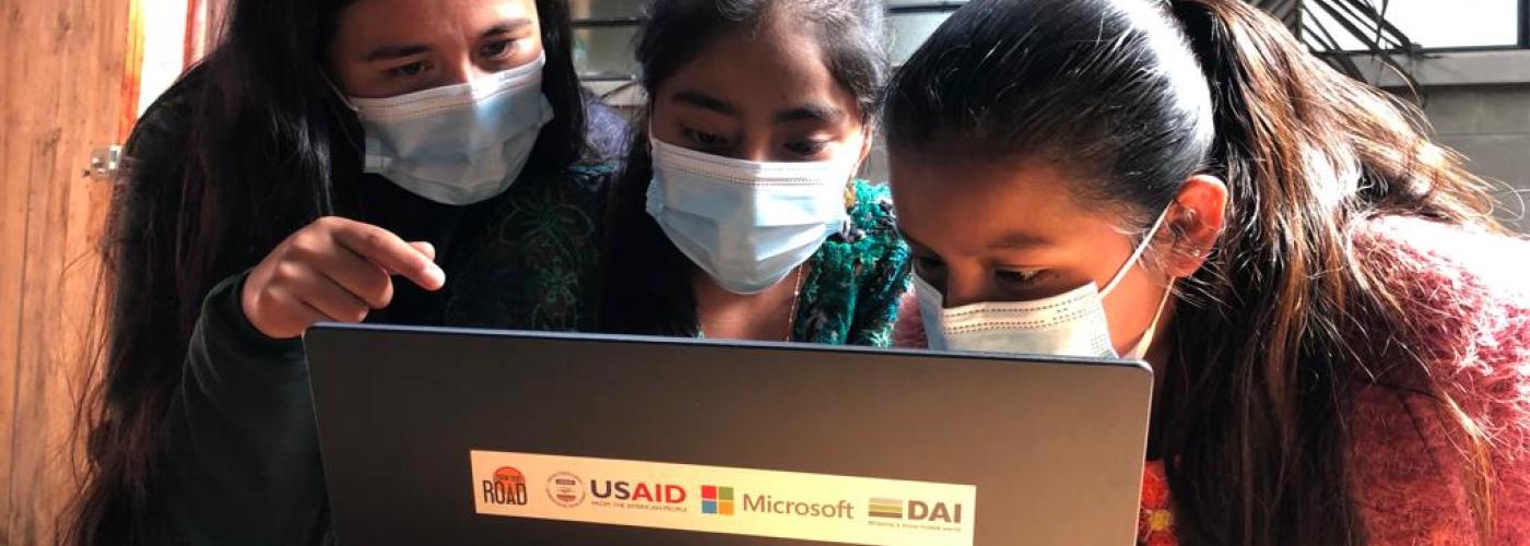 Three women wearing face masks gather around a laptop computer.