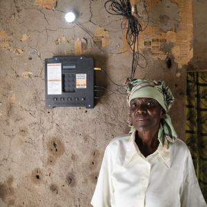 Photo: Woman standing next to an electricity generator, looking at camera. 