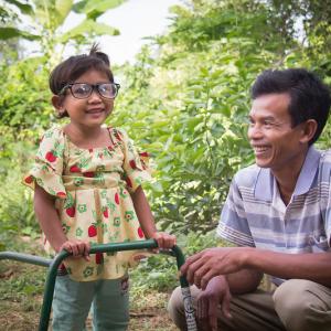 Photo: three-year-old Sreyna with glasses and a walking frame to help her see and learn to walk, standing with adult male, her father