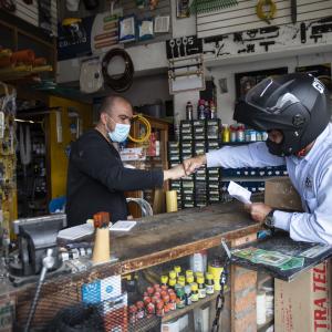 two men in a shop fist bumping. man behind the counter has a mask. man in front of the counter is wearing a motorcycle helmet 